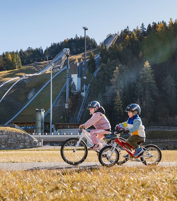 Zwei Kinder auf Fahrrädern mit Skisprungschanzen der Region Seefeld im Hintergrund.