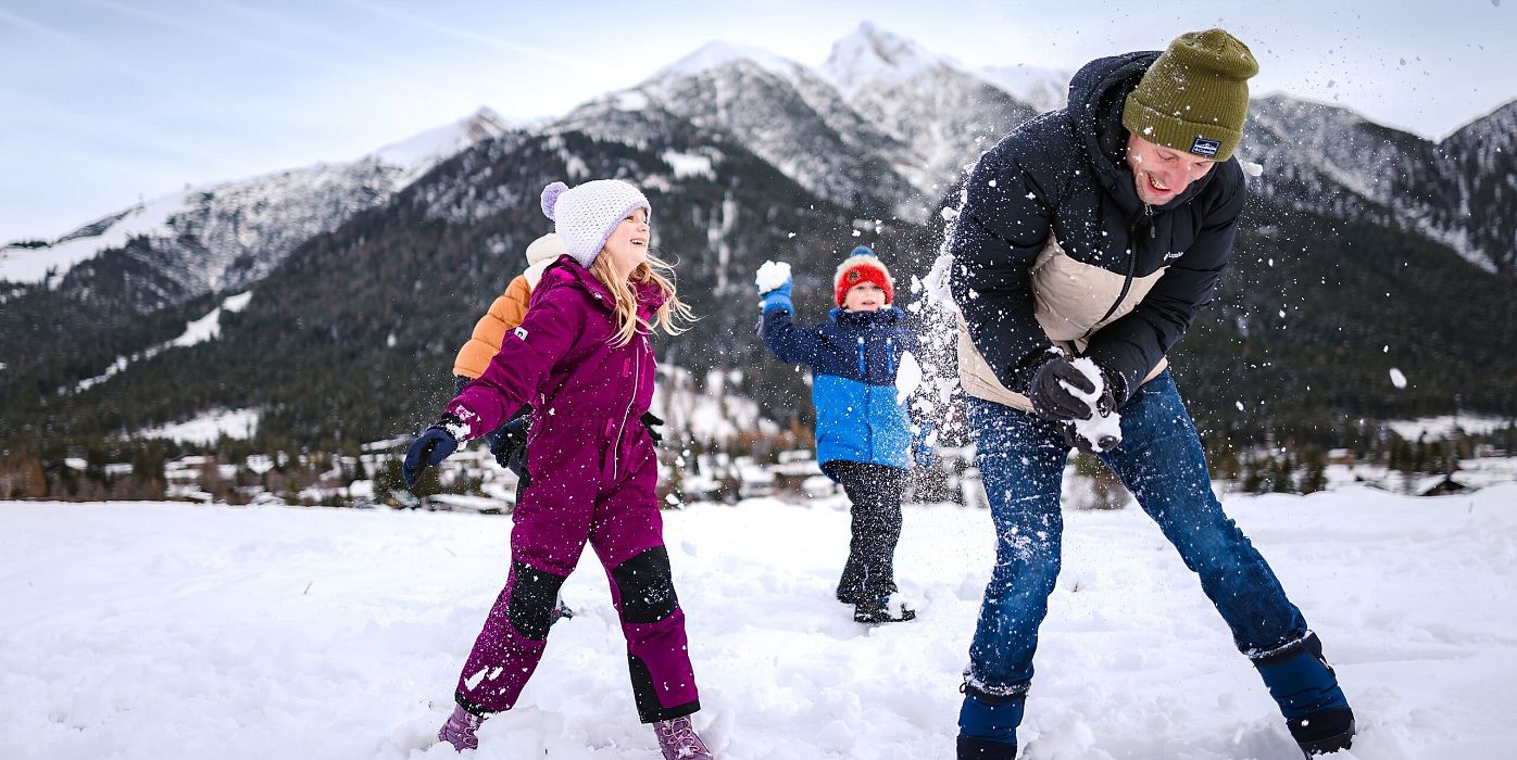 Zwei Kinder und ein Erwachsener spielen im Schnee vor bergiger Landschaft.