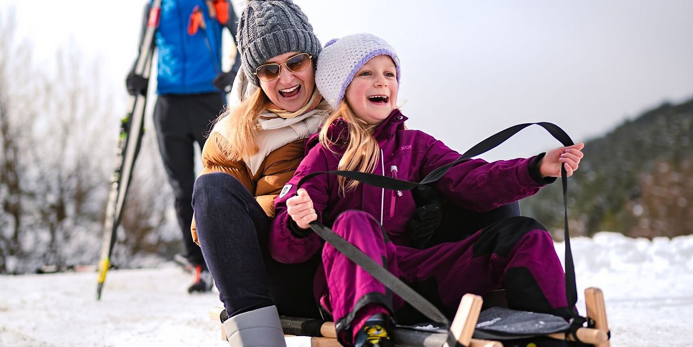 Eine Mutter und Tochter lachen auf einem Schlitten im Schnee, im Hintergrund ein Mann mit Skiern.