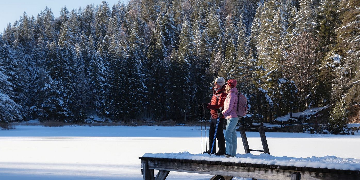 Zwei Personen stehen auf einem Holzsteg am Möserer See, umgeben von schneebedeckten Bäumen. Winterliche Berglandschaft.