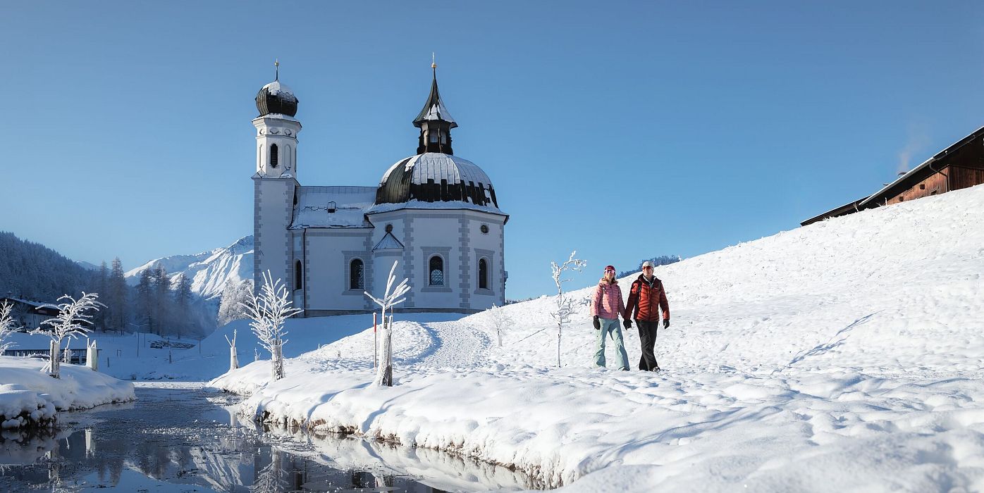 Verschneite Kirche in Winterlandschaft, Wanderer im Vordergrund. Region Seefeld. Klarer Himmel, blau.