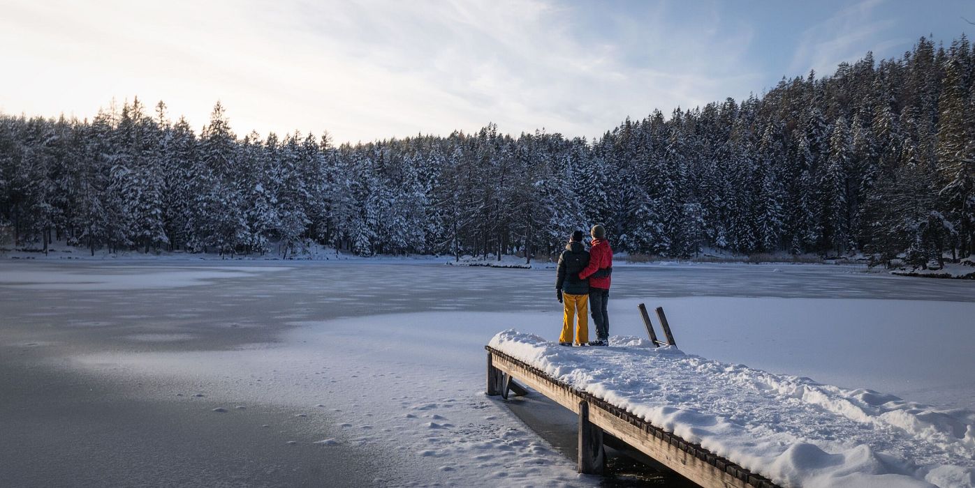 Verschneiter Steg am See in der Region Seefeld, zwei Personen, winterlicher Wald im Hintergrund.