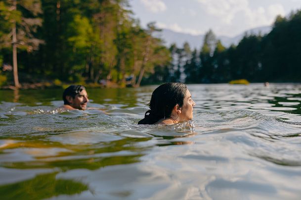 Zwei Personen schwimmen in klarem Wasser, umgeben von Bäumen und Bergen.