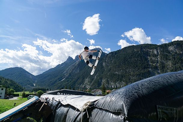 Snowboarder springt über ein große Kissen in den Bergen unter blauem Himmel.