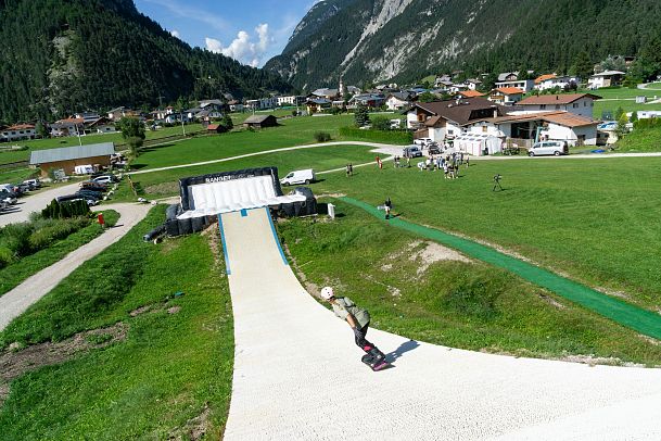Skateboarder on a ramp at a mountain resort with green hills and buildings in the background.