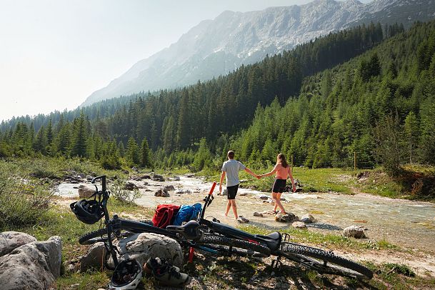 A couple walks hand in hand near a stream, with bicycles resting in the foreground.