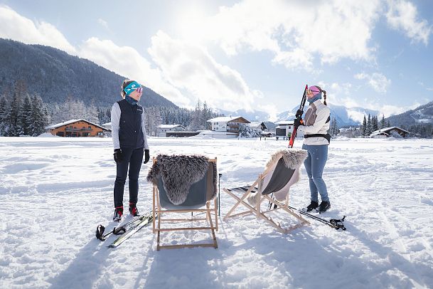 Einkehr bei der J'auszeit Hütte_zwei Frauen kommen mit Langlaufskiern bei Liegestühlen an
