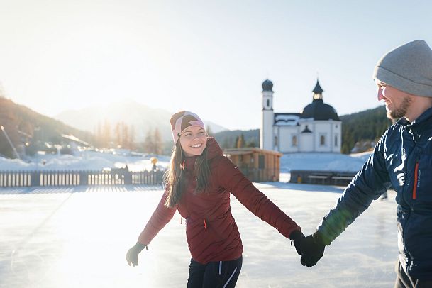 Paar beim Eislaufen auf einem Eisplatz in der Region Seefeld, mit Berglandschaft und Kirche im Hintergrund.