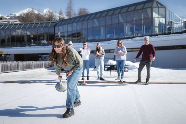 Gruppe spielt Eisstockschießen in der Region Seefeld vor modernem Gebäude auf schneebedeckter Eisfläche.
