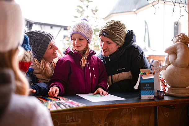 Familie malt mit Buntstiften an einem winterlichen Stand in der Region Seefeld. Schnee im Hintergrund.
