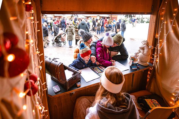 Familie auf einem Weihnachtsmarkt, schreibt im Marktstand etwas. Lichter und Dekoration schaffen eine festliche Stimmung.