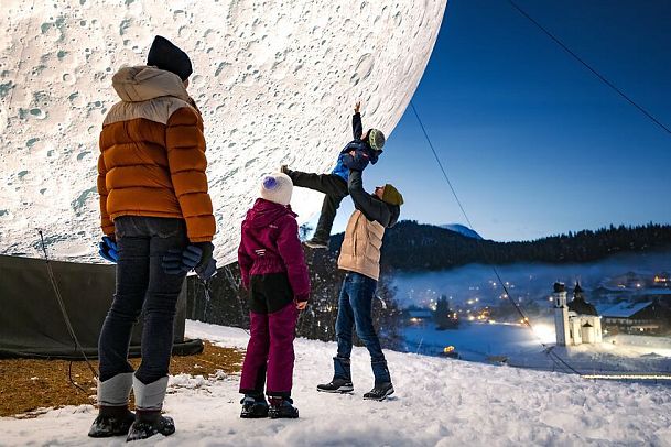 Familie vor riesigem Mondmodell im Schnee, Berge und beleuchtetes Gebäude im Hintergrund.