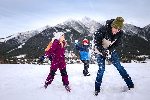 Drei Personen im Schnee vor Bergen in der Region Seefeld. Kinder werfen Schnee in die Luft. Winterkleidung, blauer Himmel.