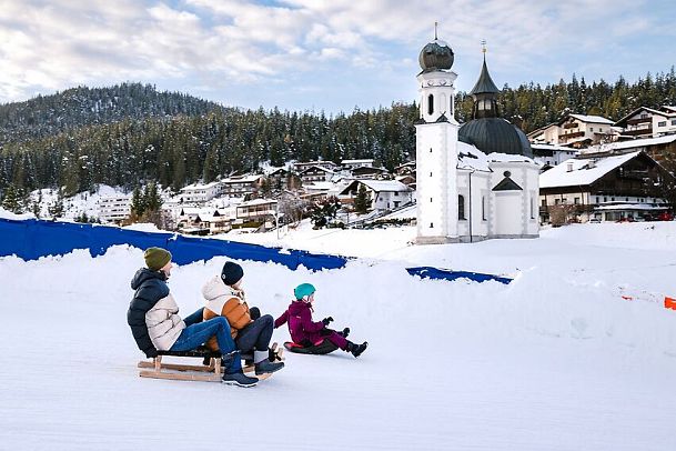 Drei Menschen rodeln vor verschneiter Kirche in der Region Seefeld. Perfekt für Winterurlaub und Rodelspaß in Tirol.