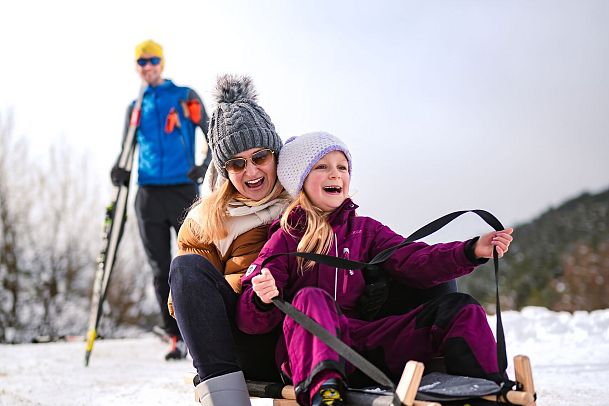 Mutter und Kind rodeln im Schnee, Skifahrer im Hintergrund, Winterlandschaft.