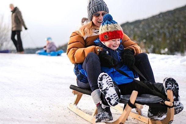 Mutter und Kind rodeln auf einem Holzschlitten einen verschneiten Hang hinunter.