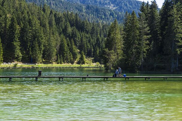 Zwei Personen stehen auf einem Steg an einem See, umgeben von Bäumen und Bergen.