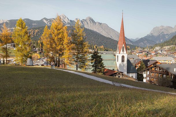 herbstspaziergang-in-seefeld-mann-und-frau-laufen-am-weg-auf-dem-pfarrhuegel-aufnahme-auf-der-distanz-mit-blick-auf-pfarrkirche-st