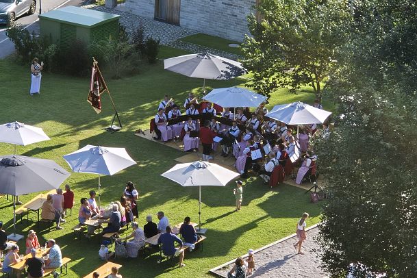 Menschen sitzen unter Sonnenschirmen bei einer Freiluftveranstaltung in einem parkähnlichen Umfeld.