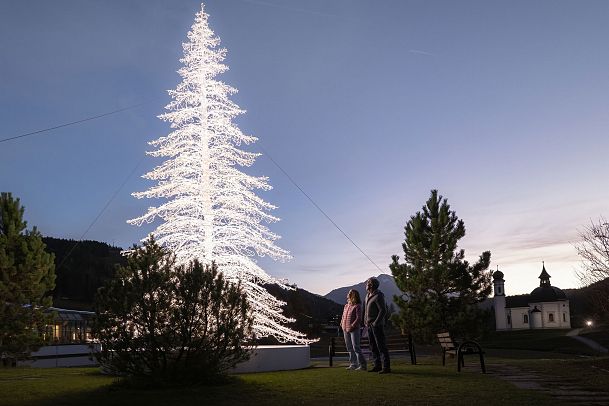Erleuchteter Weihnachtsbaum mit zwei Personen im Vordergrund unter einem Abendhimmel.