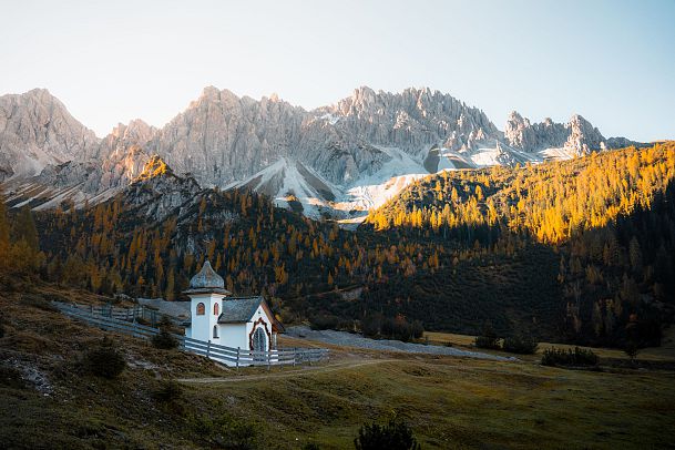 Eine kleine Kapelle in einer herbstlichen Berglandschaft mit hohen Bergen im Hintergrund.