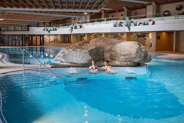 Indoor swimming pool with rock formation and two people relaxing in the water.