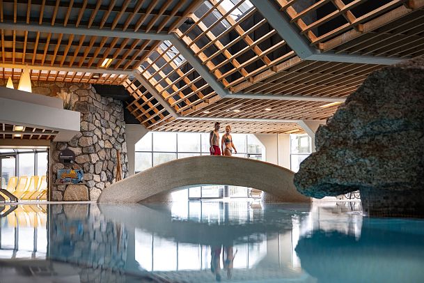 Indoor swimming pool with a bridge and two people standing, featuring natural light and stone elements.