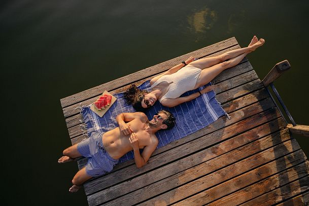 Two people lying on a dock with a towel and watermelon beside them.