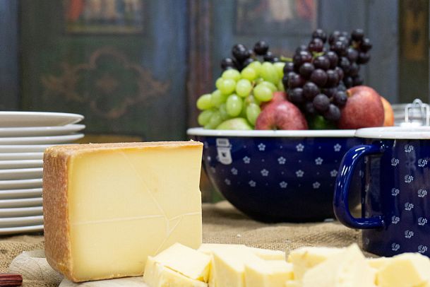A block of cheese with cut pieces, accompanied by a bowl of fruits and plates on a wooden table.