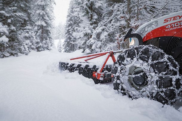 Traktor mit Schneeketten pflügt durch verschneiten Waldweg.