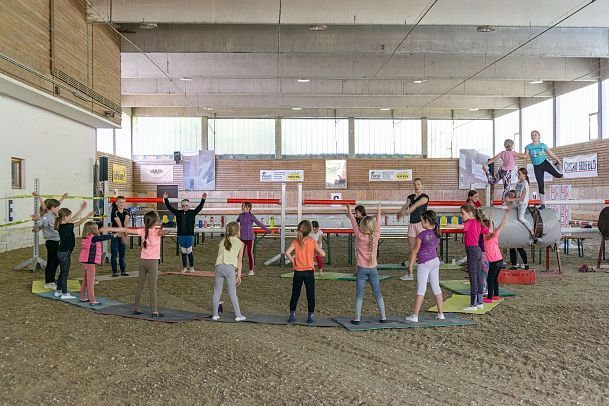 Kinder trainieren in einer Reithalle an verschiedenen Stationen und mit Hindernissen.