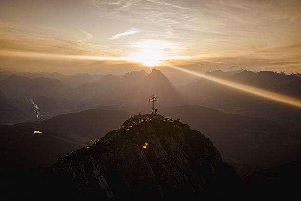 Sonnenuntergang über Bergen mit einem Kreuz auf einem Gipfel.