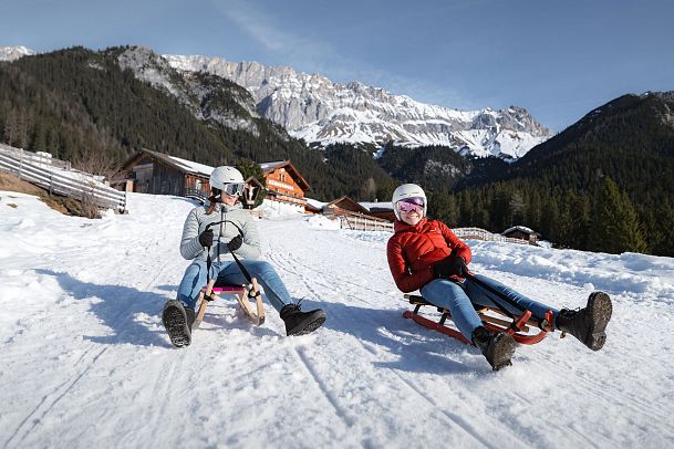 Zwei Personen rodeln einen verschneiten Hang hinunter, Berge und Hütten im Hintergrund. Wintersport in der Region Seefeld.