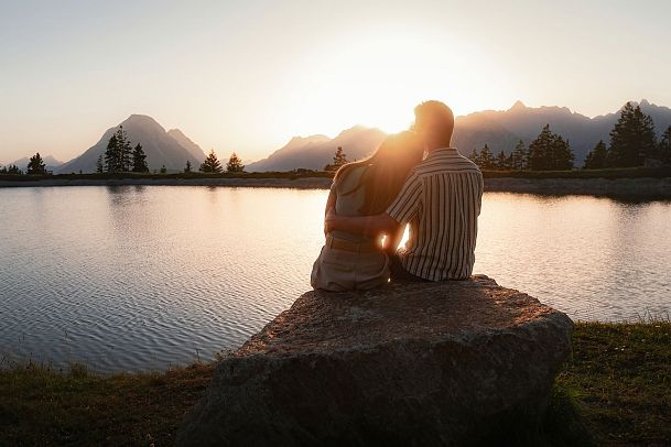 Paar sitzt auf einem Stein an einem See bei Sonnenuntergang, umgeben von Bergen.