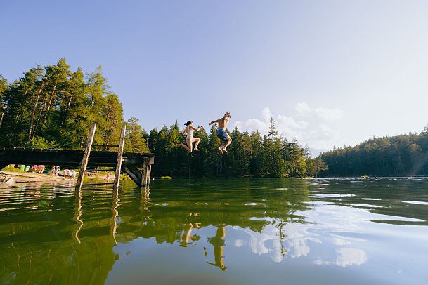 Two people jumping off a dock into a lake surrounded by trees.