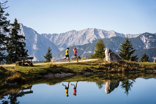 Zwei Jogger laufen am Ufer eines Bergsees in der Region Seefeld. Die Berge spiegeln sich im klaren Wasser wider.