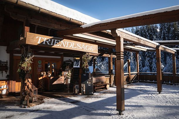 A wooden building labeled 'Triendlsäge' in a snowy landscape with benches and logs.