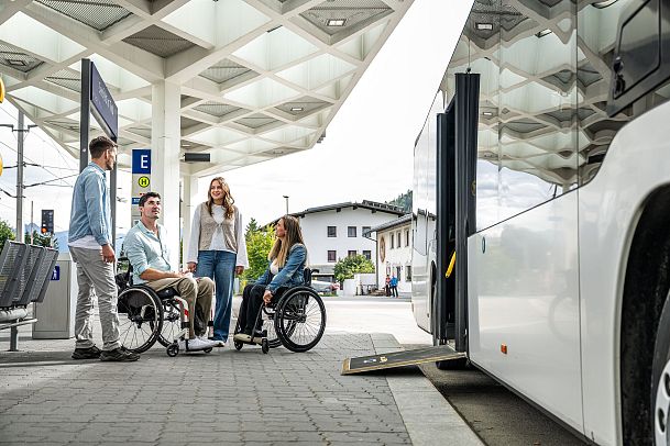 Vier Personen an einer Bushaltestelle, zwei im Rollstuhl. Der Bus hat eine Rampe für Rollstuhlfahrer.