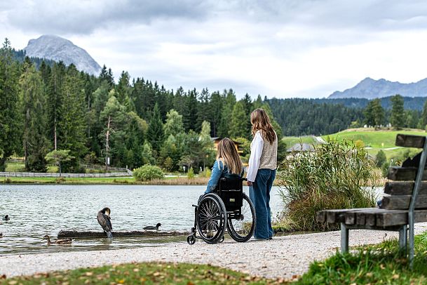 Zwei Frauen am Seeufer in der Region Seefeld, eine im Rollstuhl. Enten schwimmen im See, Berge im Hintergrund.