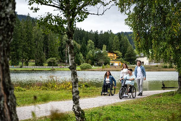 Familie am Seeweg in der Region Seefeld, zwei Personen in Rollstühlen, malerische Umgebung mit Bäumen und bewölktem Himmel.