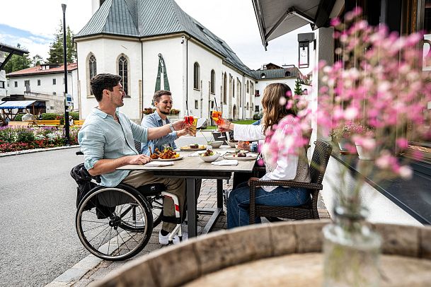 Drei Personen essen draußen in einem Bergdorf Nähe der Region Seefeld, mit einer Kirche im Hintergrund. Eine Person sitzt im Rollstuhl.