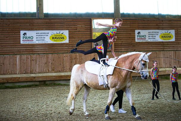 Voltigierer auf einem Pferd in einem Reitstall, Mädchen steht auf dem Pferd, andere in bunten Kostümen.