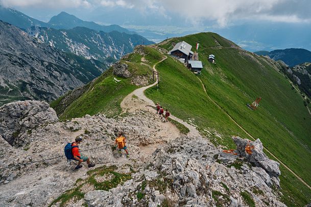 Eine Berglandschaft mit Wanderern auf einem Pfad und einer Hütte im Hintergrund.