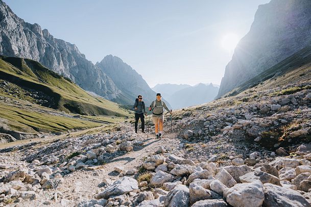 Zwei Personen wandern auf einem unebenen Pfad zwischen hohen Bergen.