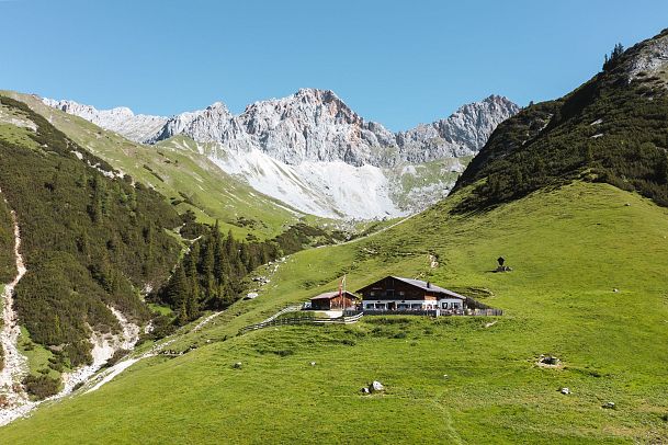 A wooden chalet in green meadows with mountains in the background under a clear blue sky.
