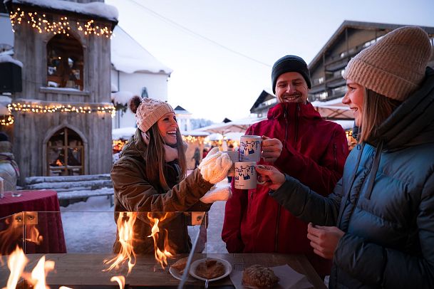 Weihnachtsmarkt mit Menschen drei Freunde stoßen mit Glühwein an Glockenturm links Feuer zu sehen