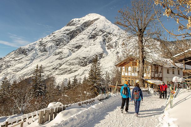 People walking on a snowy path with mountains and wooden houses in the background.
