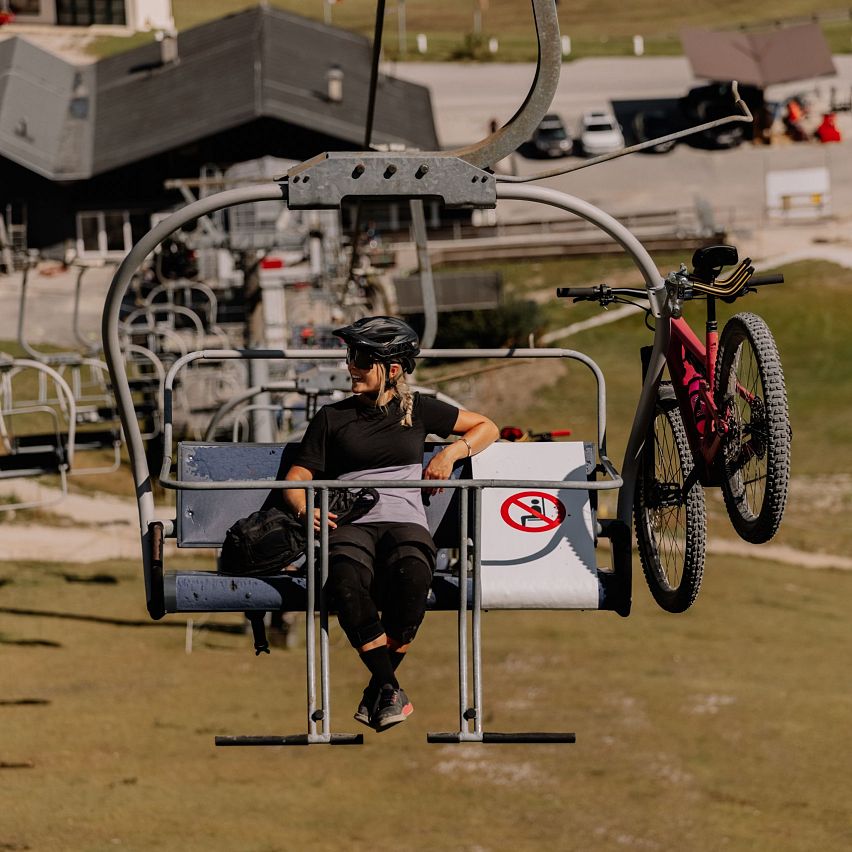 Fahrradfahrer mit Mountainbike auf Sessellift. Im Hintergrund Berge und Hütte.