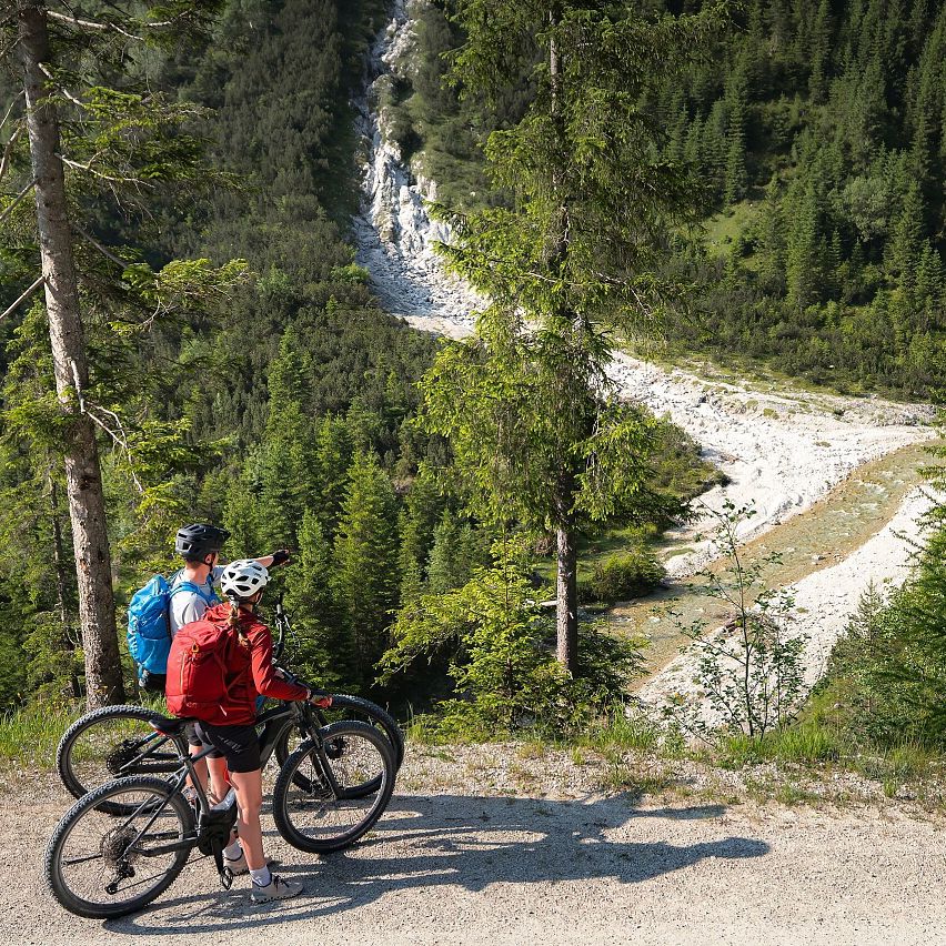 E-Biken im Gaistal im Sommer_zwei Biker_innen auf Forstweg blicken hinab auf Ache
