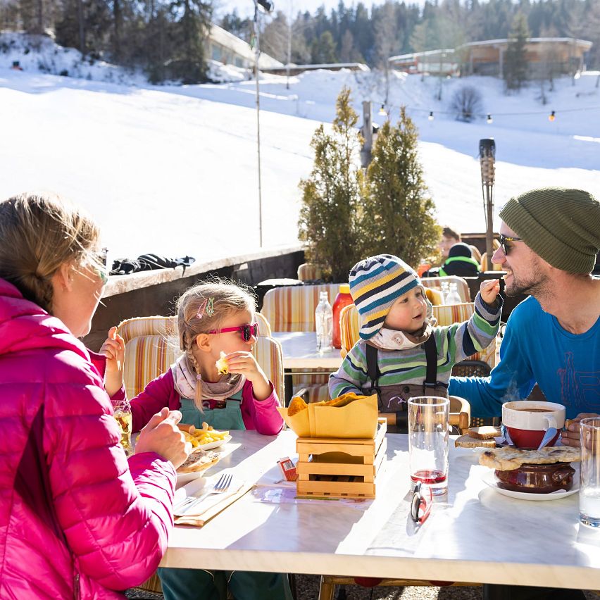 Familie sitzt bei Sonnenschein auf einer Terrasse und isst auf einem Tisch mit Blick auf die verschneite Berglandschaft.